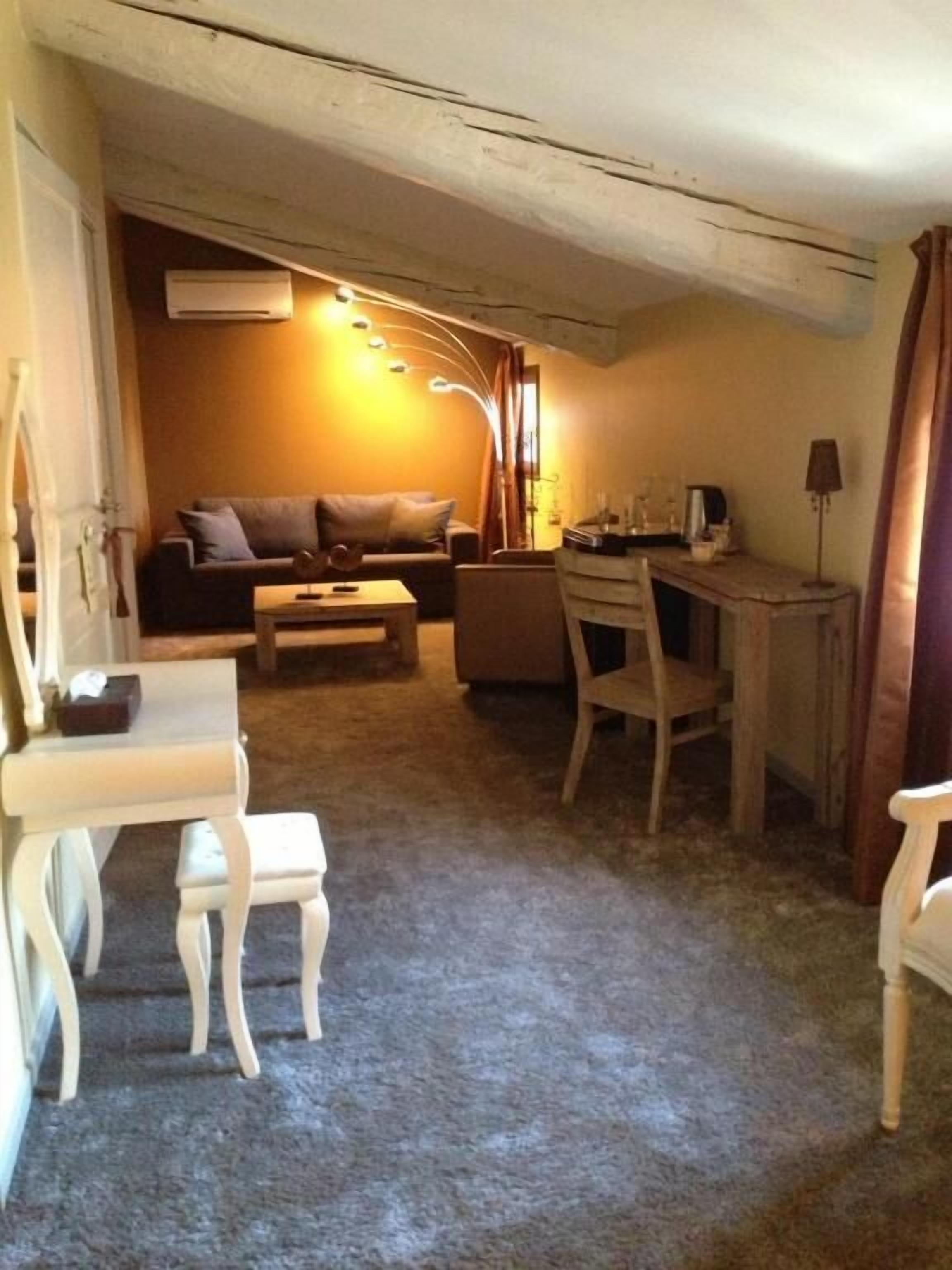 Un espacio habitable compacto con un sofá, una mesita, un escritorio y una silla. Hay una lámpara en el escritorio y un equipo de aire acondicionado en la pared.