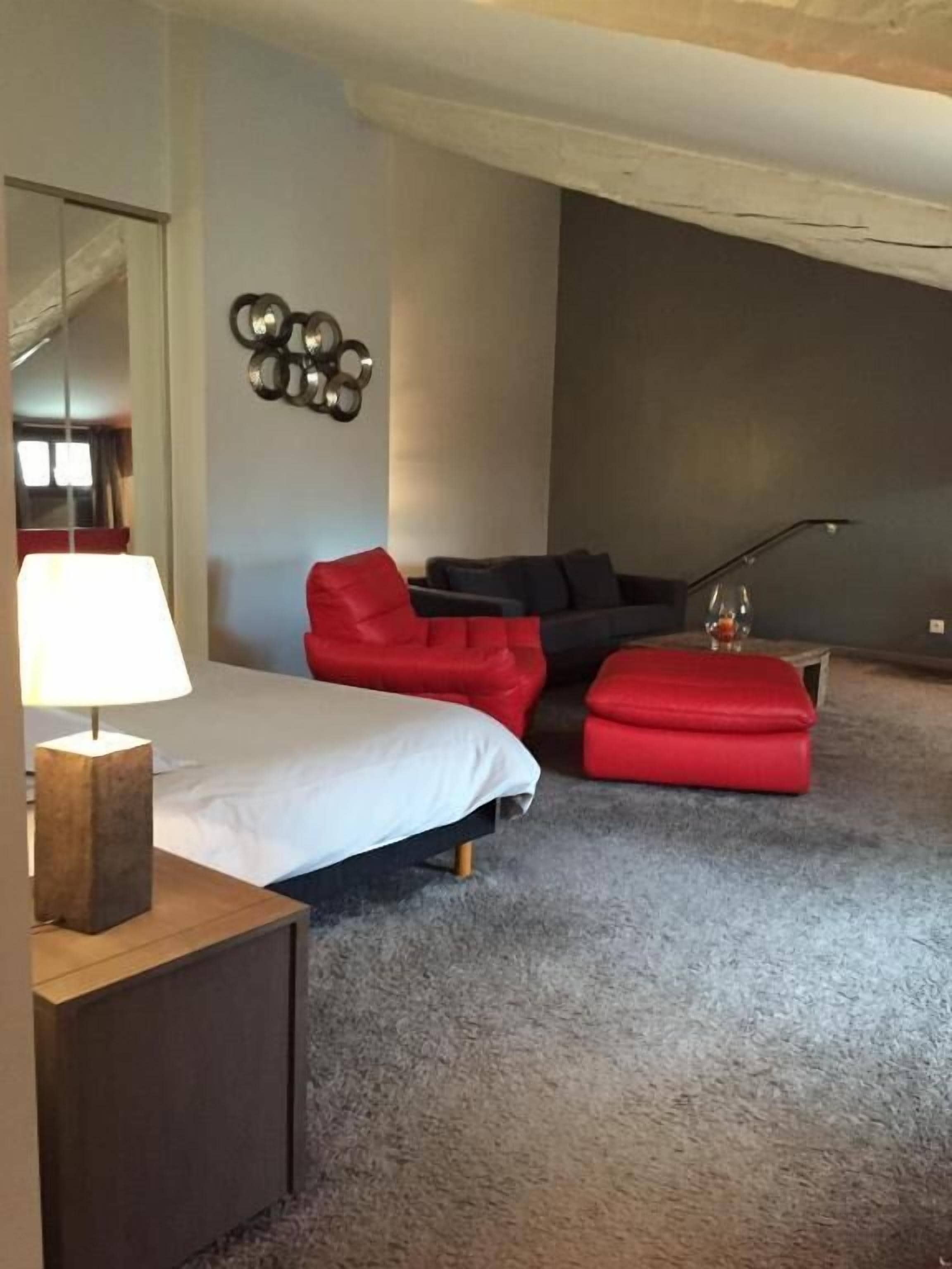 A bedroom with a bed, a red sofa, a red ottoman, a lamp, and a wall decoration.