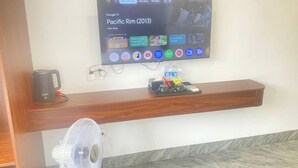 Zona de recepción de un hotel con una televisión montada que muestra un programa, un mostrador de madera, una tetera, un ventilador y un estante con agua embotellada y otros artículos.