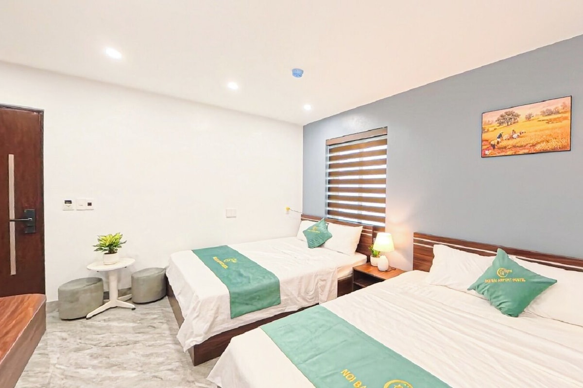 Habitación de hotel con dos camas, una mesita con una planta, una ventana con persianas y un cuadro en la pared.