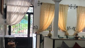 Ein Wohnzimmer mit einer Couch, Sesseln, einem Couchtisch und einem gemusterten Teppich. Es gibt ein Fenster mit Vorhängen, eine dekorative Decke und einen Blick ins Freie.