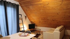 Um quarto acolhedor com teto de madeira, um sofá branco, uma mesa de jantar com cadeiras e uma televisão.