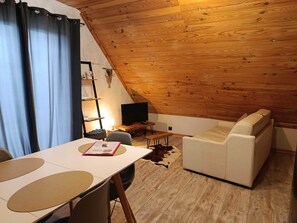 Ein gemütliches Zimmer mit Holzbalkendecke, einem weißen Sofa, einem Esstisch mit Stühlen und einem Fernseher.