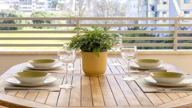 Una mesa de madera redonda con una maceta amarilla de plantas verdes, servida con cuencos verdes, platos blancos y copas de vino.