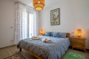 A bedroom with a bed, two bedside lamps, a wooden nightstand, a green rug, and a wall-mounted artwork. - Casa Eolo. Appartamento con terrazza e box privato. (Alghero)