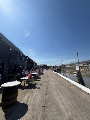 An outdoor dining area with tables and chairs, a building with signage, and a body of water in the background. - Stylish 3-Bed Home | Garden, Parking & Near Quay (Kent)