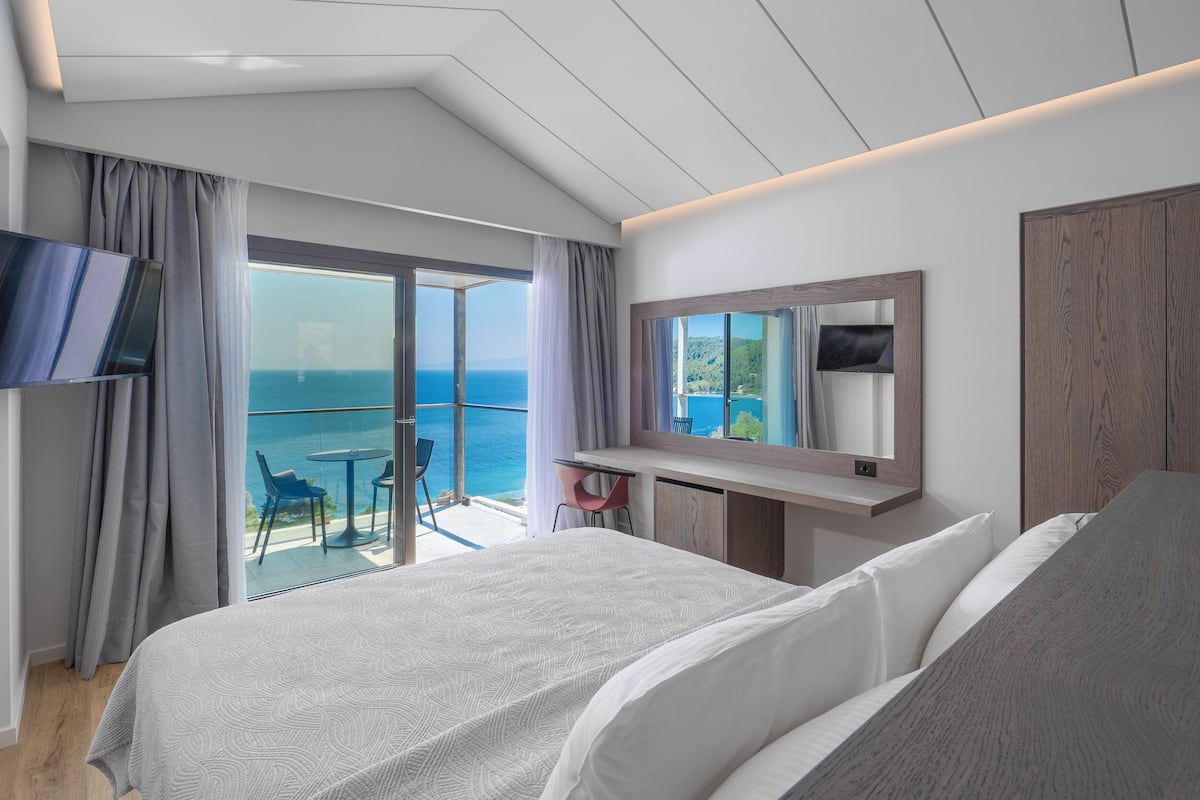 A hotel room with a bed, a TV, a desk, and a balcony with a view of the sea.