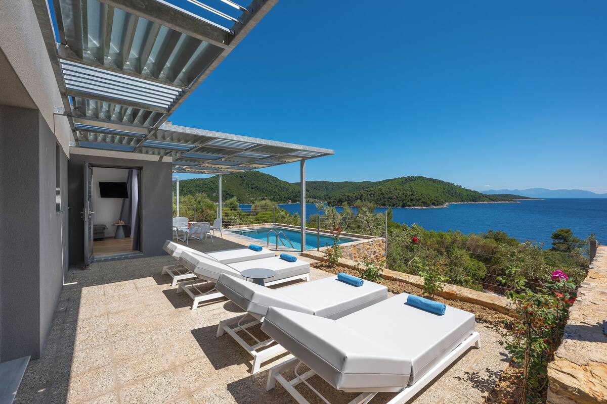 A poolside area with sun loungers, a covered seating area, and a view of the sea and hills.