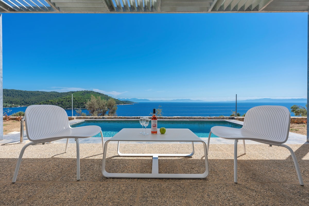 A terrace with two white chairs and a table, overlooking a pool and a scenic view of the sea and hills.