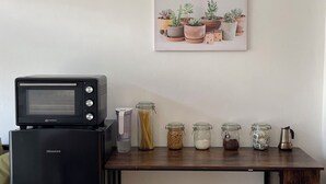 Eine Küchenzeile mit einem schwarzen Kühlschrank, einer Mikrowelle und einer Kaffeemaschine auf einem Holztisch. Es stehen Gläser mit Lebensmitteln auf dem Tisch, und an der Wand hängt ein gerahmtes Bild von Pflanzen.