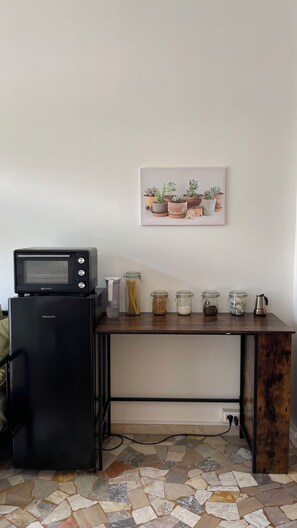 Eine Küchenzeile mit einem schwarzen Kühlschrank, einer Mikrowelle und einer Kaffeemaschine auf einem Holztisch. Es stehen Gläser mit Lebensmitteln auf dem Tisch, und an der Wand hängt ein gerahmtes Bild von Pflanzen.