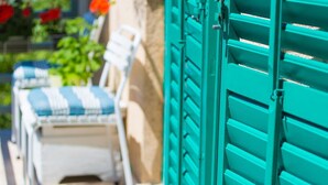 A sunlit outdoor seating area with a white chair and a blue and white striped cushion, a green shutter door, and a plant with red flowers in the background. - Apartment Bijenica - surrounded by lush greenery (Vrbnik)