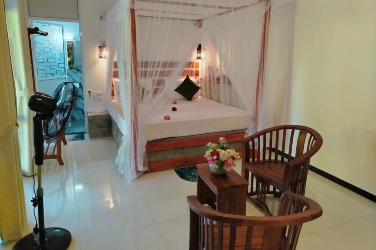 Una habitación con una cama con dosel, una silla de madera, una mesita con un florero y un ventilador.