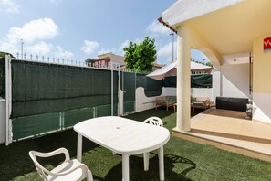 Um pátio com mesa e cadeiras brancas, área de estar coberta e cerca verde.