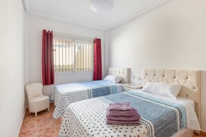 Une chambre avec deux lits, une chaise, une fenêtre avec des barreaux et un rideau rouge.
