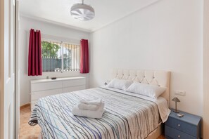 Quarto com cama, mesa de cabeceira, cômoda, janela com cortinas e luminária no teto.