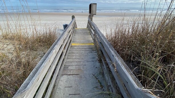 Ein hölzerner Steg führt zu einem Strand, im Hintergrund ist eine Holzkonstruktion zu sehen.