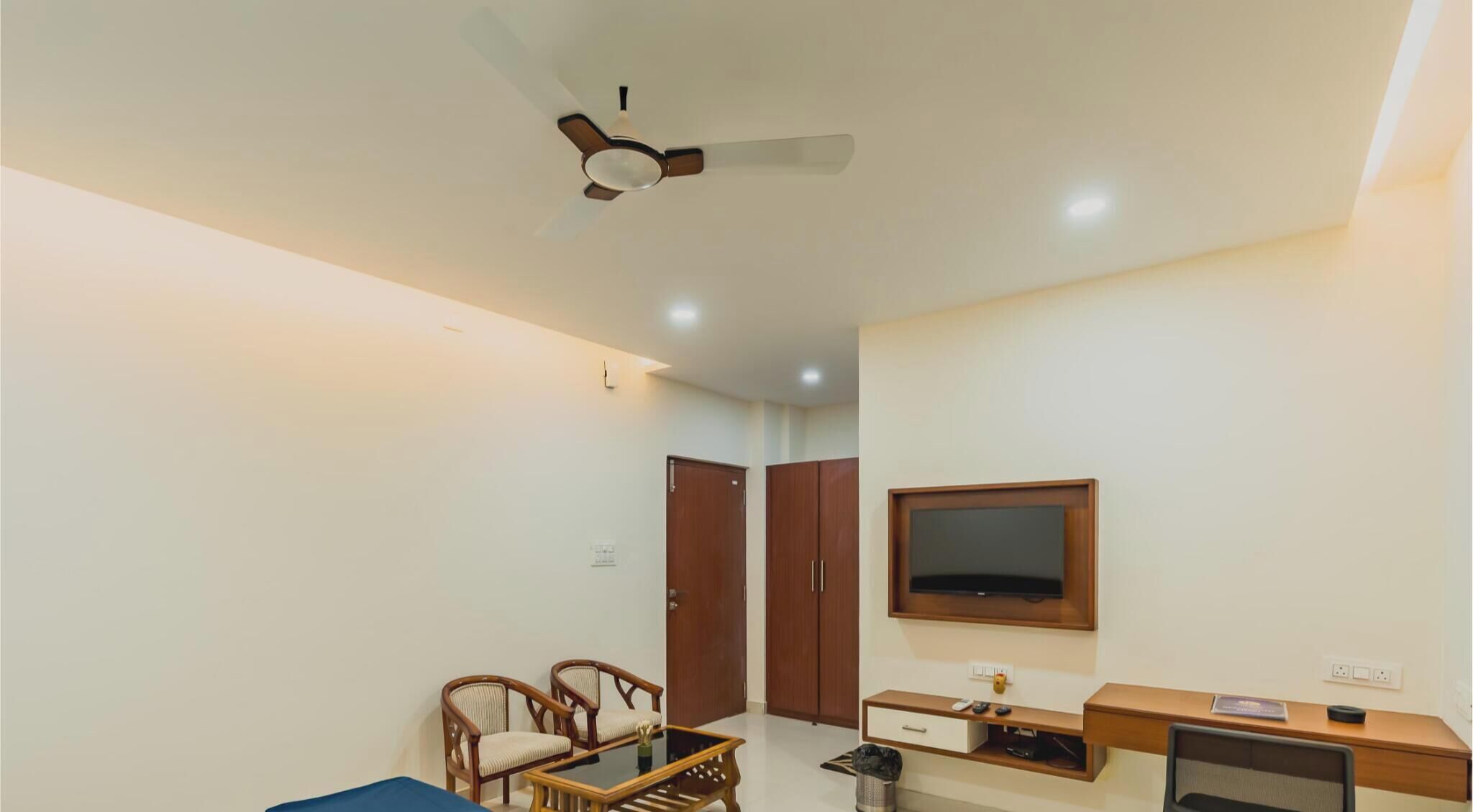Una sala de estar con ventilador de techo, televisor montado en la pared, mueble de madera, escritorio y dos sillas.