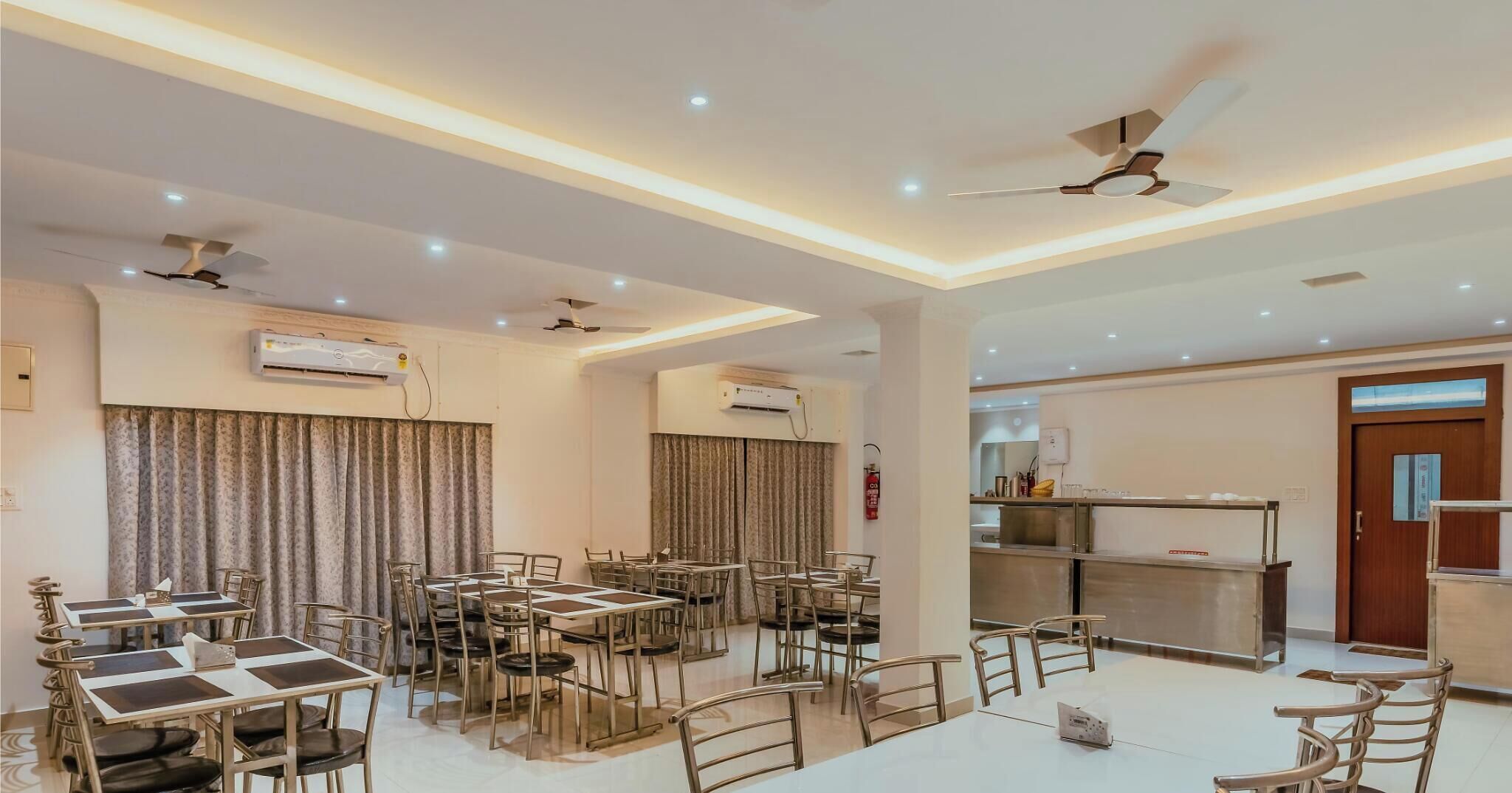 Un comedor de hotel con varias mesas y sillas, ventiladores de techo, unidades de aire acondicionado y una cocina visible a través de un panel de vidrio.