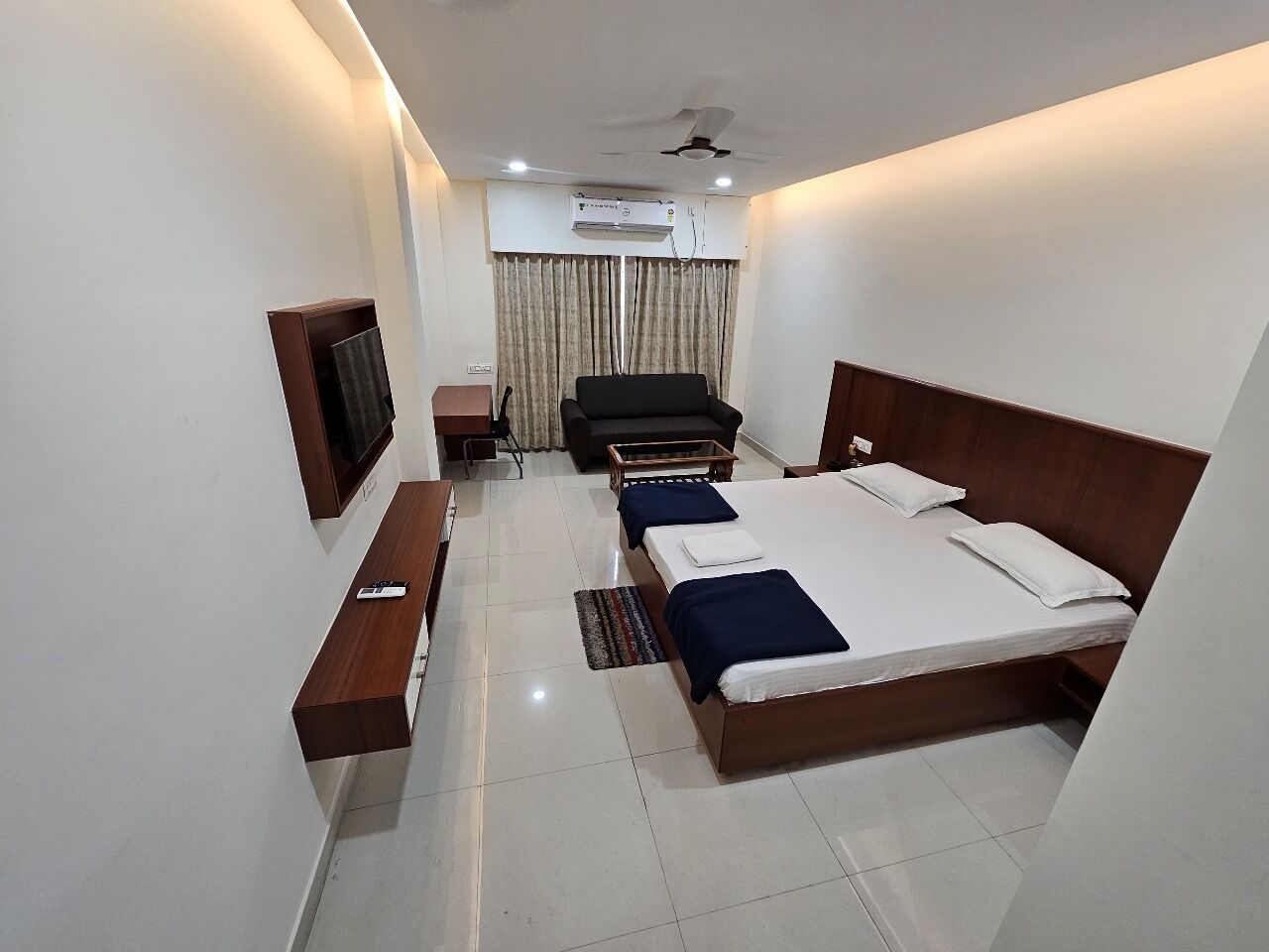 A hotel room with a bed, a desk, a chair, a TV, and an air conditioning unit.