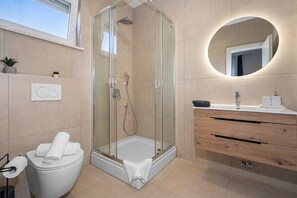 A modern bathroom with a glass-enclosed shower, a toilet with a towel, a wooden vanity, and a round mirror. - Villa Vallis (Sveta Nedelja)