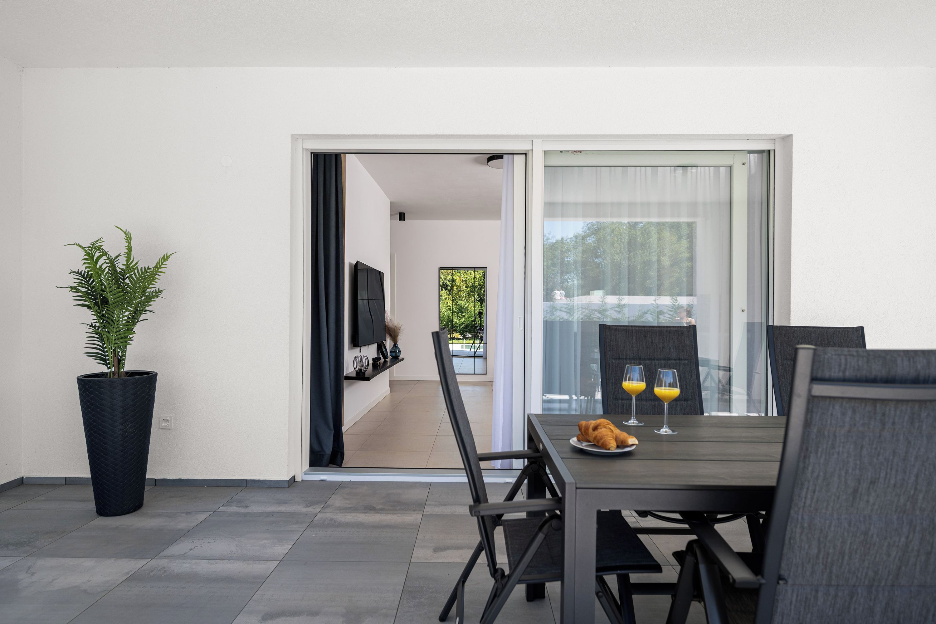 A modern dining area with a table set for two, a potted plant, and a view through a glass door.