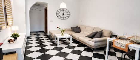 Ein modernes Wohnzimmer mit Schachbrettmuster-Boden, einer Couch, einem Couchtisch, einem Esstisch mit Stühlen, einer Wanduhr und einer Pendelleuchte.
