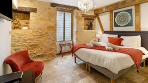 Quarto com parede de pedra, cama com roupa de cama branca e vermelha, poltrona vermelha e um candelabro.