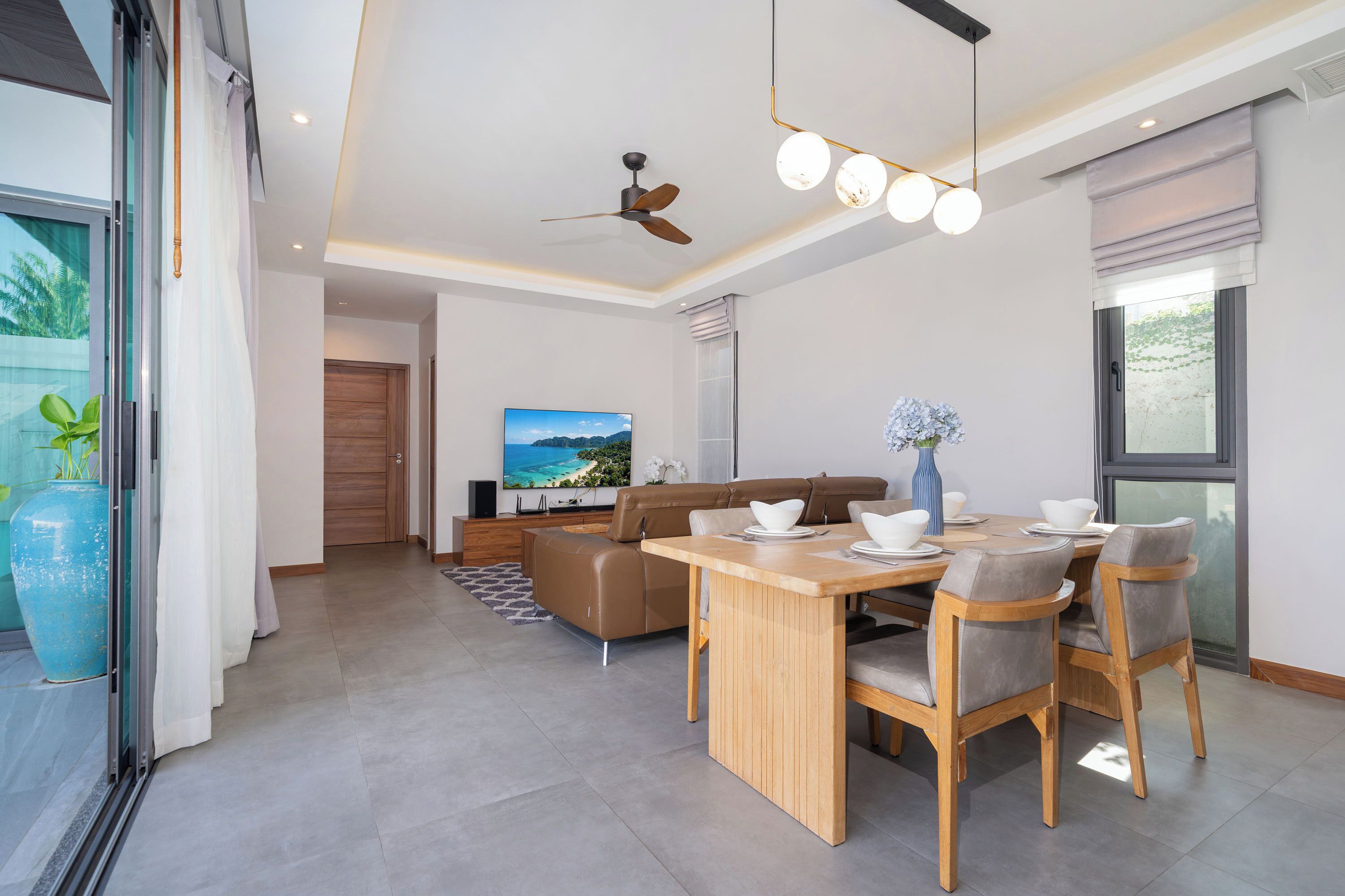 A modern living room with a sofa, dining table, chairs, a ceiling fan, and a TV.