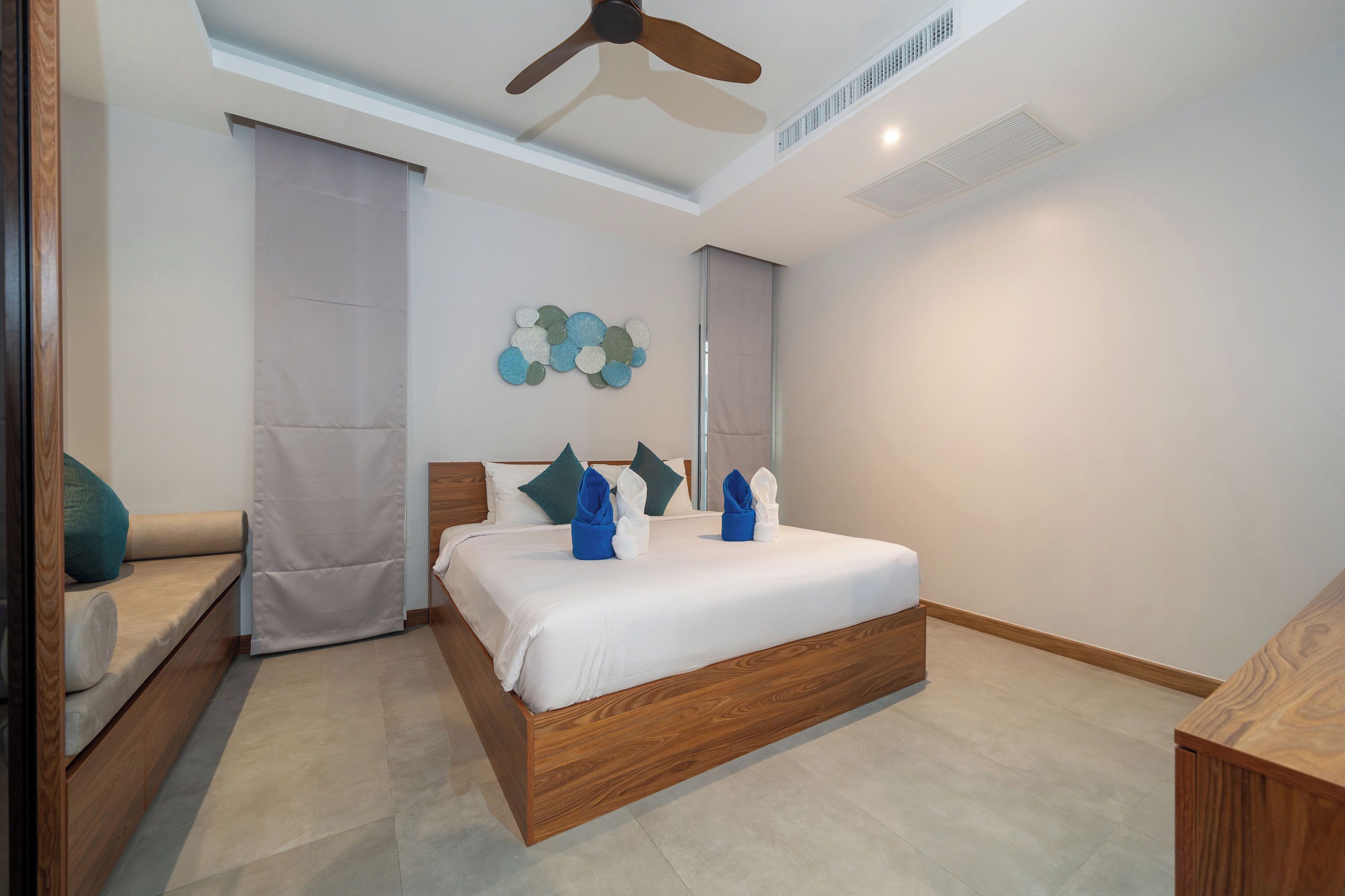A modern hotel room with a wooden bed, a ceiling fan, a wall-mounted artwork, and a seating area with a bench and cushions.