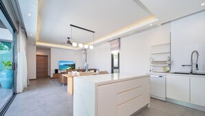 A modern kitchen with a central island, a dining area with a sofa and table, and a large window with curtains. - Nicky Serene 3BR Pool Villa (Choeng Thale)