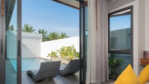 A modern living room with a glass door leading to a pool area, featuring striped lounge chairs and a yellow pillow. - Nicky Serene 3BR Pool Villa (Choeng Thale)