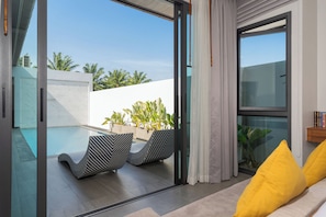 A modern living room with a glass door leading to a pool area, featuring striped lounge chairs and a yellow pillow. - Nicky Serene 3BR Pool Villa (Choeng Thale)