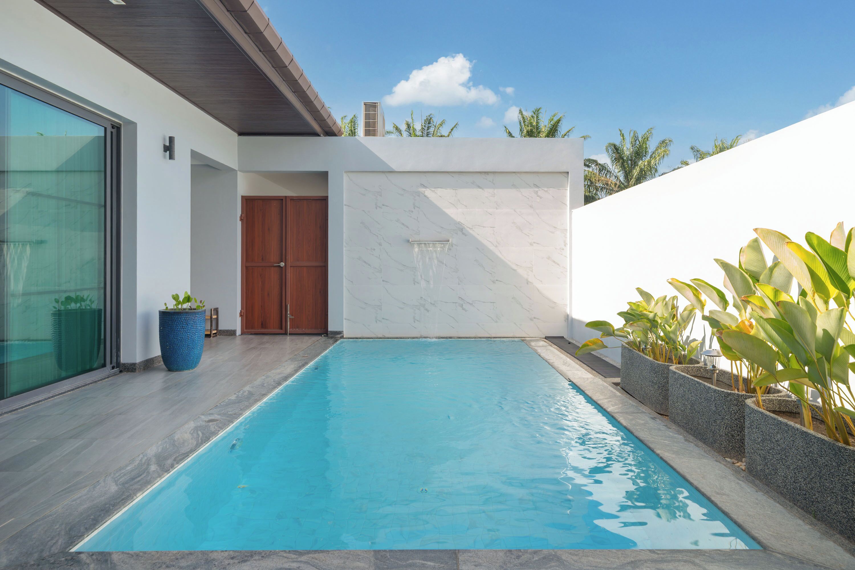 A modern pool area with a clear blue pool, a wooden door, and a white wall with a marble pattern.