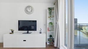 A modern living room with a flat-screen TV, a white entertainment unit, a wall clock, a plant shelf, and a view of a river and buildings. - Riverside Apartment Greenwich 2Bed Near O2 (London)