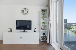 Ein modernes Wohnzimmer mit einem Flachbildfernseher, einer weißen Unterhaltungsecke, einer Wanduhr, einem Pflanzenregal und Blick auf einen Fluss und Gebäude.