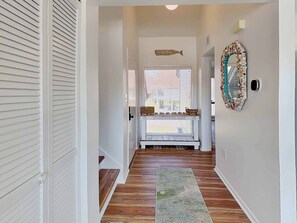 A hallway with a wooden floor, a bench with baskets, a decorative mirror, and a door leading to a room. - Heron Marsh Villa 30, Newly Renovated on Lake! (Pawleys Island)