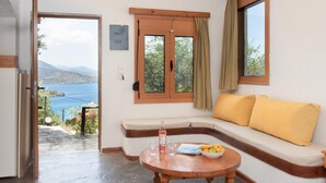 Una habitación con vista al mar, un sofá con cojines amarillos, una mesa con una botella y vasos, y una puerta que da al exterior.