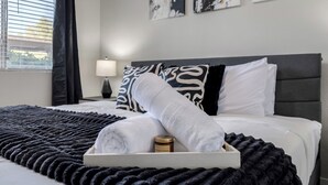 A neatly made bed with a rolled white towel, a jar of French vanilla, and a black and white patterned pillow.