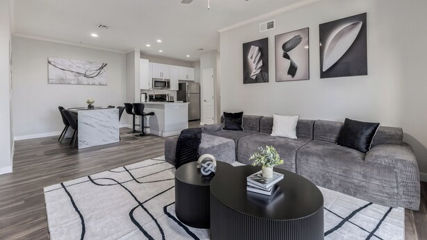 A modern living room with a grey sofa, black ottoman, and a coffee table. There is a kitchen in the background with white cabinets and a microwave.