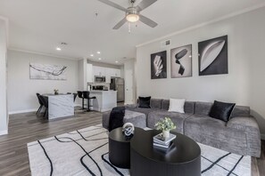A modern living room with a grey sofa, black ottoman, and a coffee table. There is a kitchen in the background with white cabinets and a microwave. - Luxury 2-bedroom Condo with AC in lovely Scottsdale (Scottsdale)