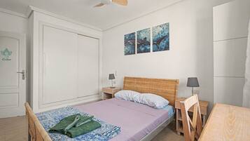 Un dormitorio con cama, mesita de noche, una silla, ventilador de techo y una obra de arte en la pared.