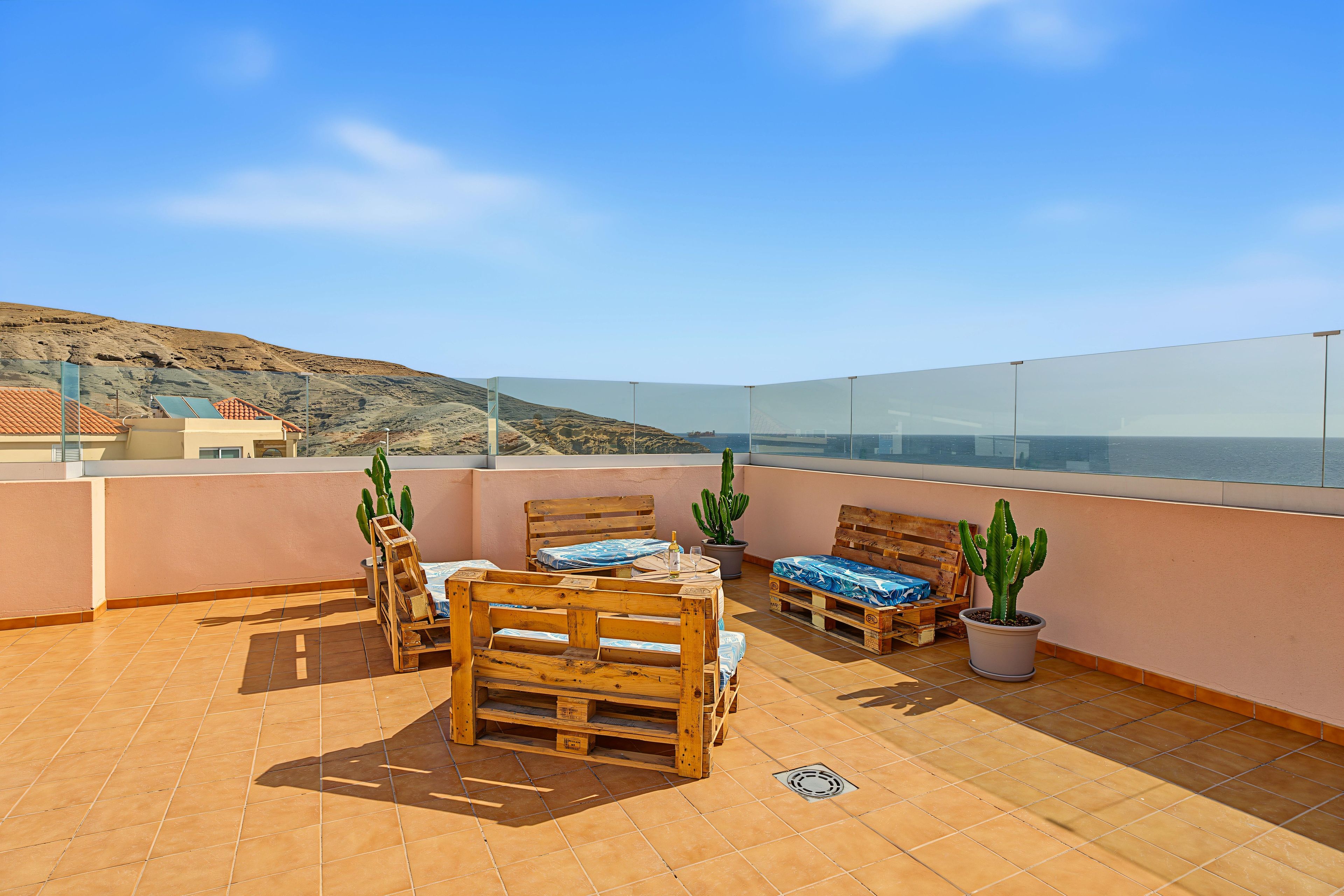 Una terraza en la azotea con muebles de madera, cactus en macetas y una vista despejada del mar y colinas lejanas.