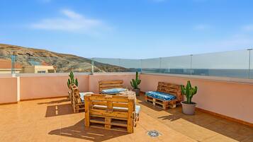Una terraza en la azotea con muebles de madera, cactus en macetas y una vista despejada del mar y colinas lejanas.