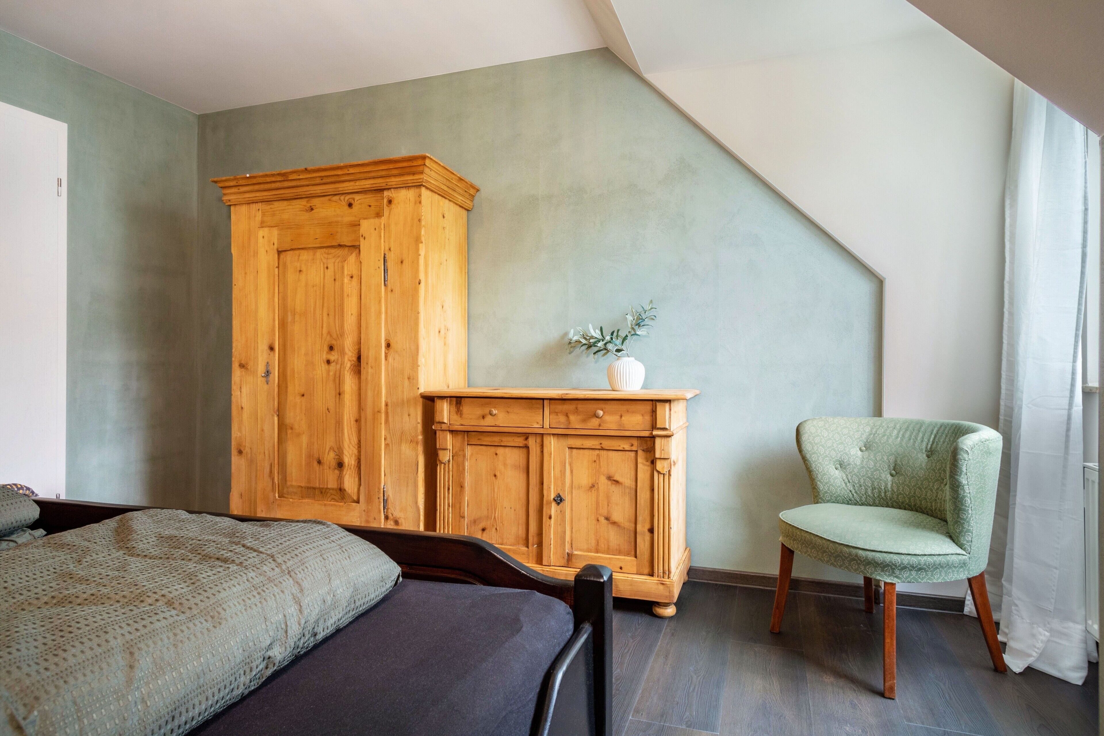 Quarto com cama, guarda-roupa de madeira, poltrona verde e um pequeno vaso branco com uma planta.
