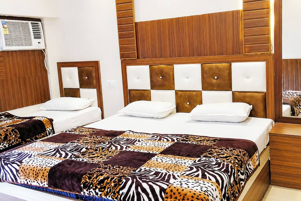 A hotel room with a large bed, a wooden headboard, a wall-mounted air conditioner, and a small bedside table.