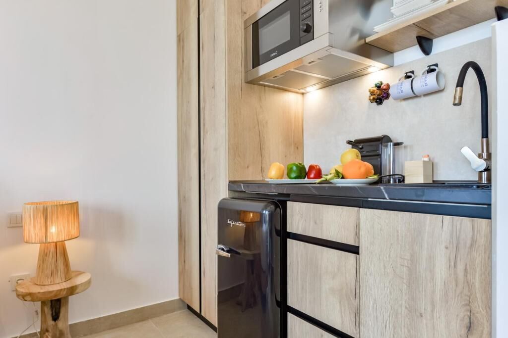 A compact kitchen with a microwave, a small refrigerator, and a sink. There are fruits on the counter, and a lamp is on a small table.