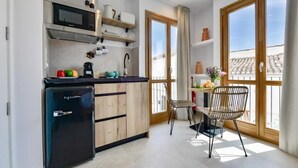 Una cocina compacta con refrigerador, microondas y gabinetes de madera. Hay un comedor con una silla y una mesa, y ventanas grandes que ofrecen vista a la ciudad.