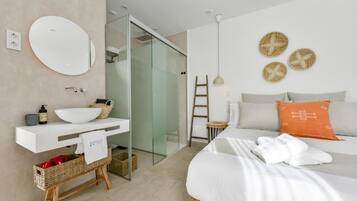 Una habitación de hotel moderna con cabina de ducha de vidrio, un lavamanos blanco con cesta, una cama con almohadas blancas y naranja, y un espejo montado en la pared.