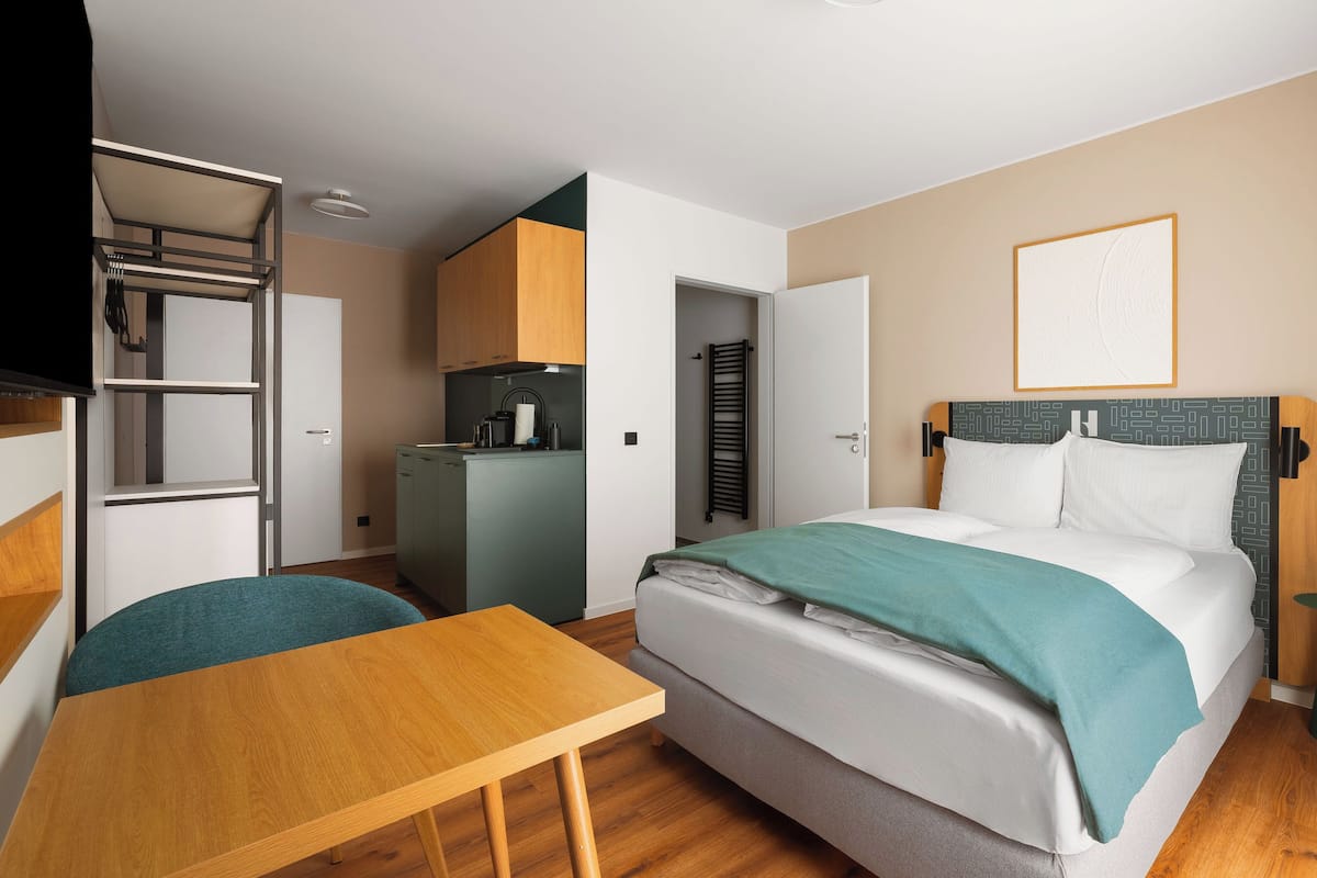 A compact hotel room with a bed, a small desk, a kitchenette, and a wardrobe.